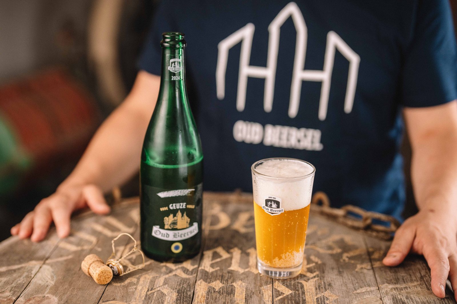 A bottle of Oude Geuze Oud Beersel next to a poured glass, on a wooden cask head
